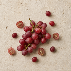 A cluster of red grapes with a few loose and halved grapes scattered on a textured beige stone surface.