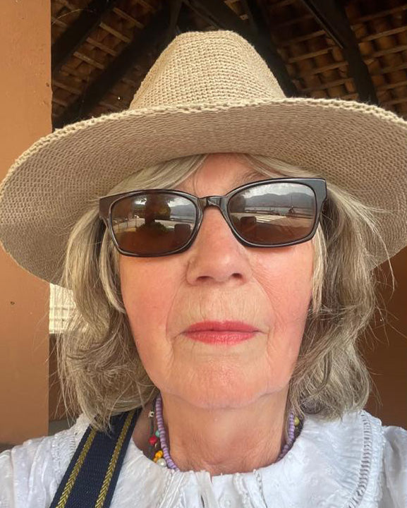 A close-up portrait of a woman wearing a beige woven sun hat, dark sunglasses, and a white top, photographed outdoors.