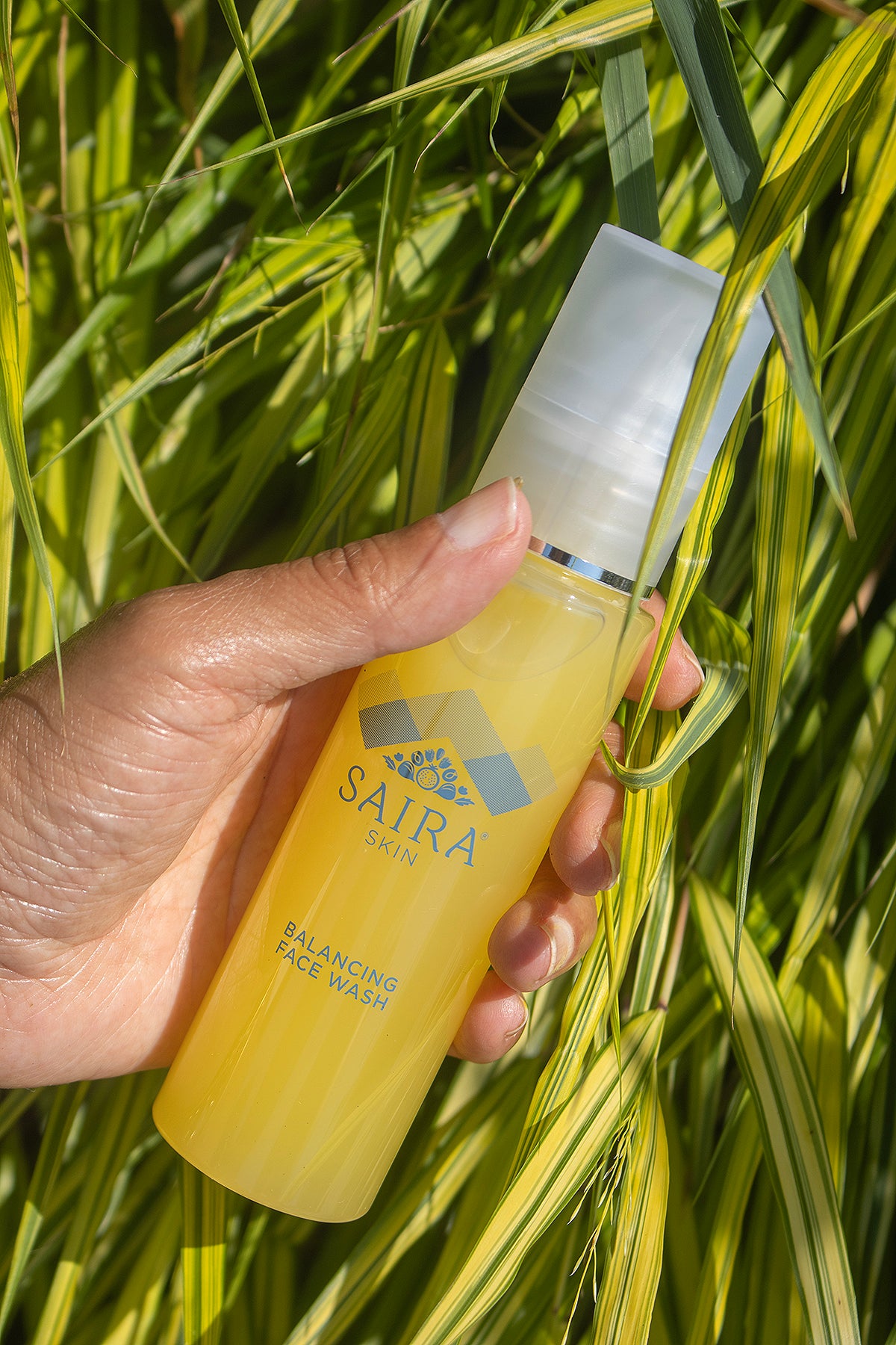 A hand holding a bottle of SAIRA Balancing Facial Wash against a background of green and yellow grass.