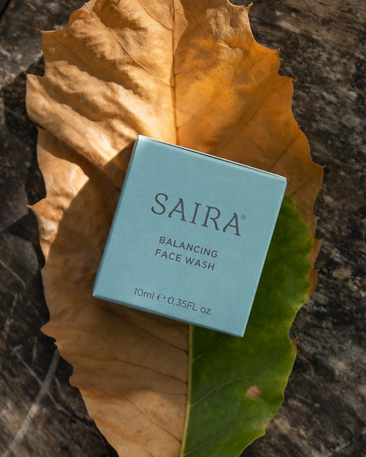 A light blue square box of SAIRA Balancing Face Wash placed on a large dried brown leaf over a dark textured surface.
