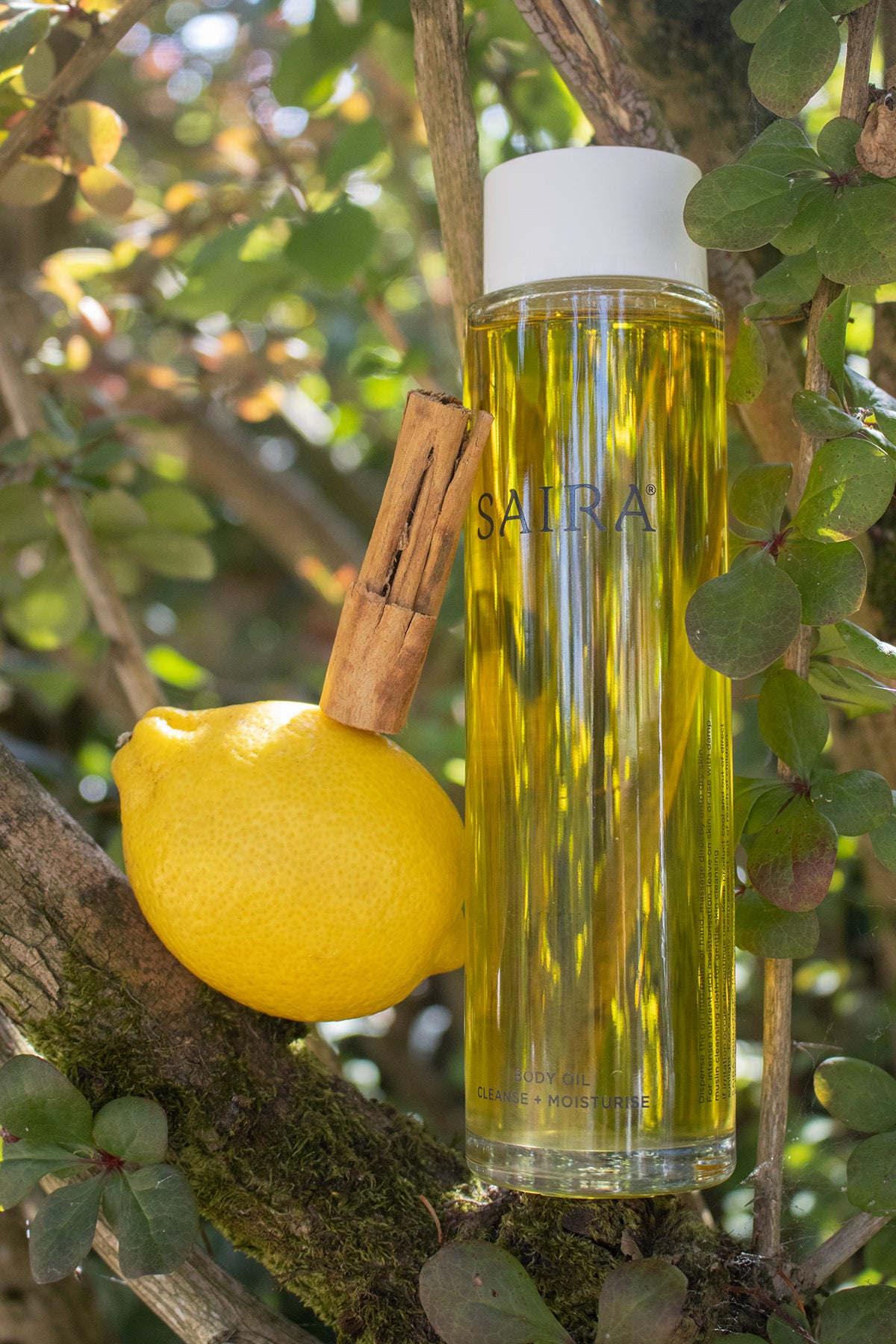 A clear cylindrical bottle of SAIRA Body Oil with a white cap, standing on a tree branch outdoors with green leaves around it. A yellow lemon and a cinnamon stick are placed beside the bottle as styled props.