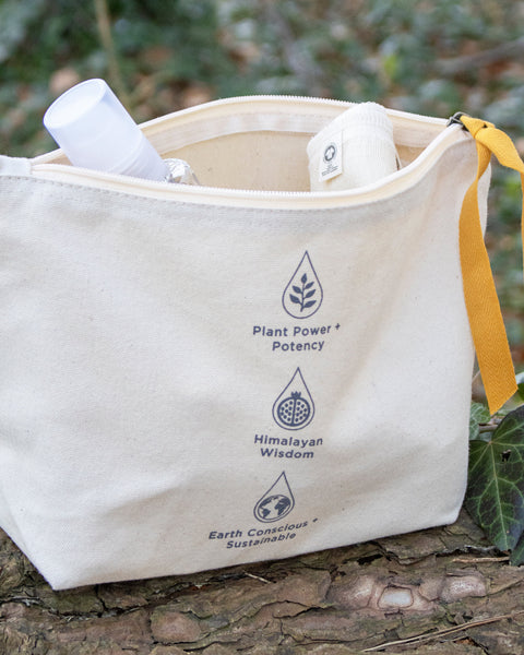 An off-white canvas cosmetic pouch with a yellow zipper pull and strap, featuring printed text and icons for Plant Power + Potency, Himalayan Wisdom, and Earth Conscious + Sustainable. The pouch is shown open, containing skincare items, resting on a tree trunk outdoors with green foliage in the background.