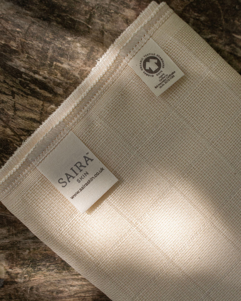 A close-up of a cream-coloured muslin cloth, laid on a rustic wooden surface. The fabric features two labels: a larger one reading 'SAIRA SKIN' and a smaller one indicating 'ORGANIC COTTON'.