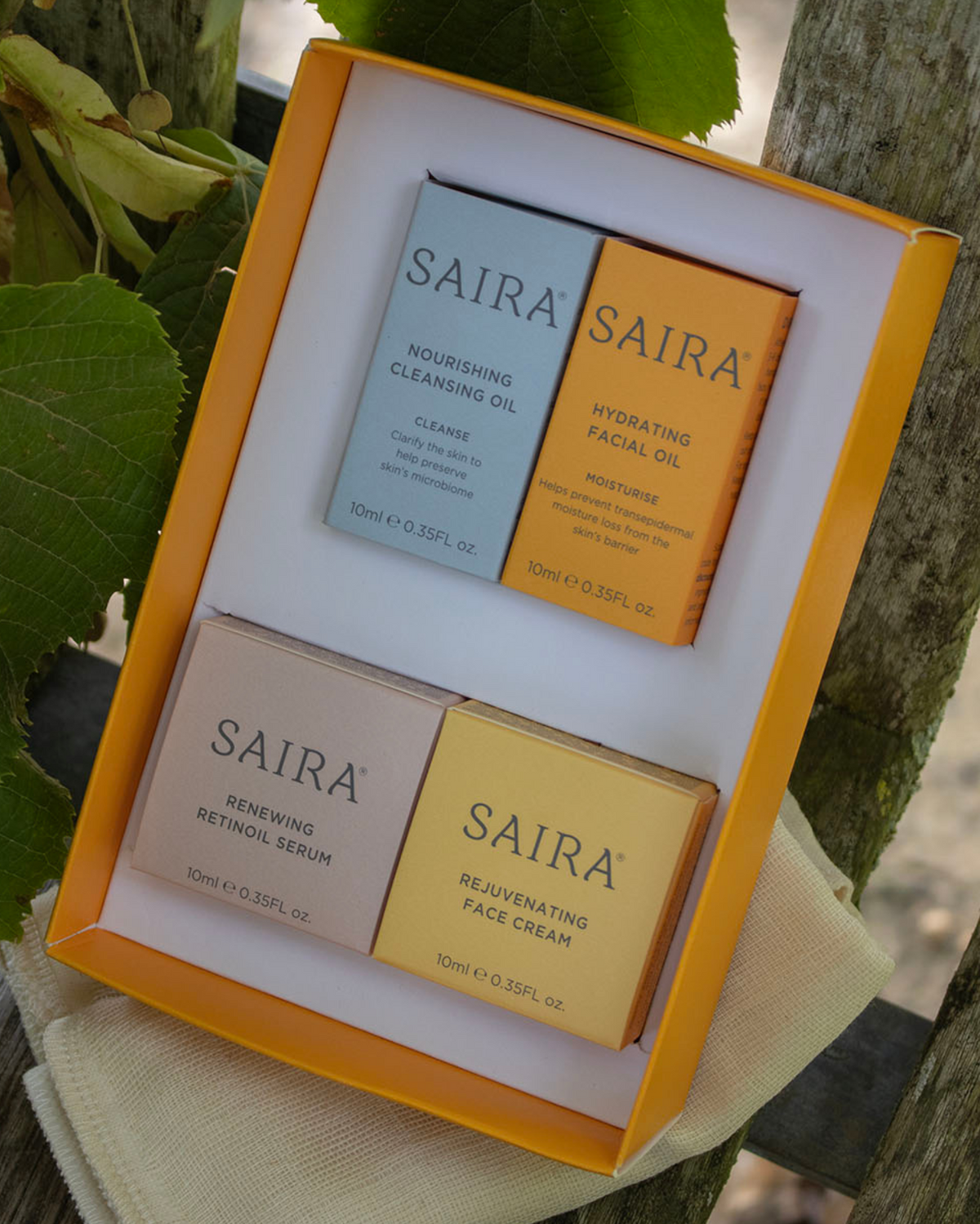 A boxed set of four SAIRA skincare products arranged in a bright orange tray, photographed outdoors on a wooden surface with leaves and branches around it. The set includes cleansing oil, hydrating facial oil, renewing retinol serum, and rejuvenating face cream.