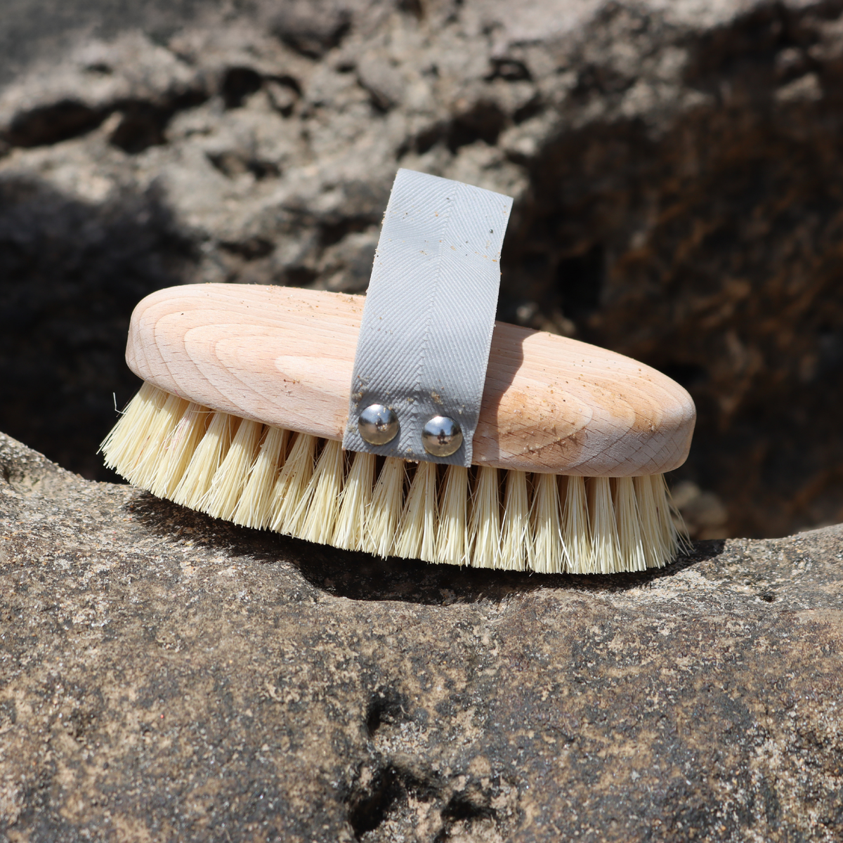 A natural wood grooming brush with cream-colored bristles and a gray fabric wrist strap, resting on a textured rock outdoors.