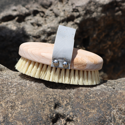 A natural wood grooming brush with cream-colored bristles and a gray fabric wrist strap, resting on a textured rock outdoors.