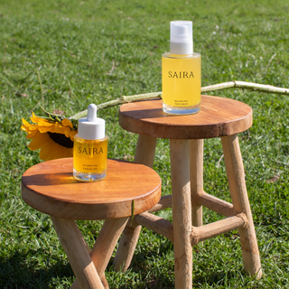 Two clear cosmetic bottles from the brand Saira, one with a dropper and one with a pump, filled with yellow oil, displayed on two small wooden stools on a green grass lawn with a sunflower prop.
