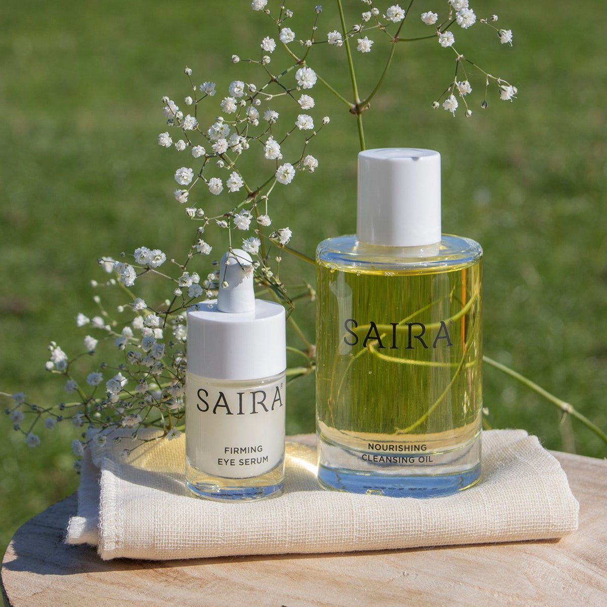 Two Saira skincare bottles, a smaller white dropper bottle labeled Firming Eye Serum and a larger clear bottle with yellow oil and a white cap labelled Nourishing Cleansing Oil, arranged on a folded cream cloth over a wooden surface with baby's breath flowers against a soft green outdoor background.