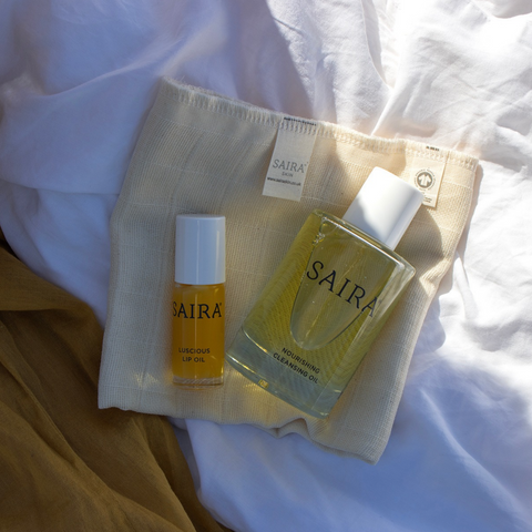 SAIRA skincare set including a cleansing oil bottle and a lip oil bottle, arranged on a cream mesh pouch over white draped fabric.