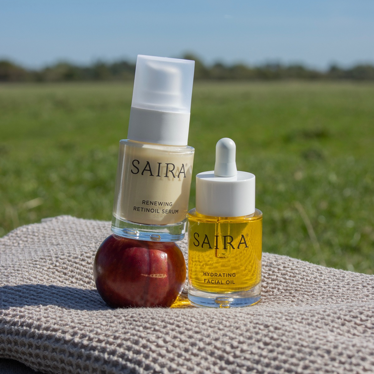 Two skincare products from Saira, a retinol serum and a facial oil, displayed on a textured beige blanket outdoors with a green field and blue sky in the background. The products are styled with a red apple on the left.