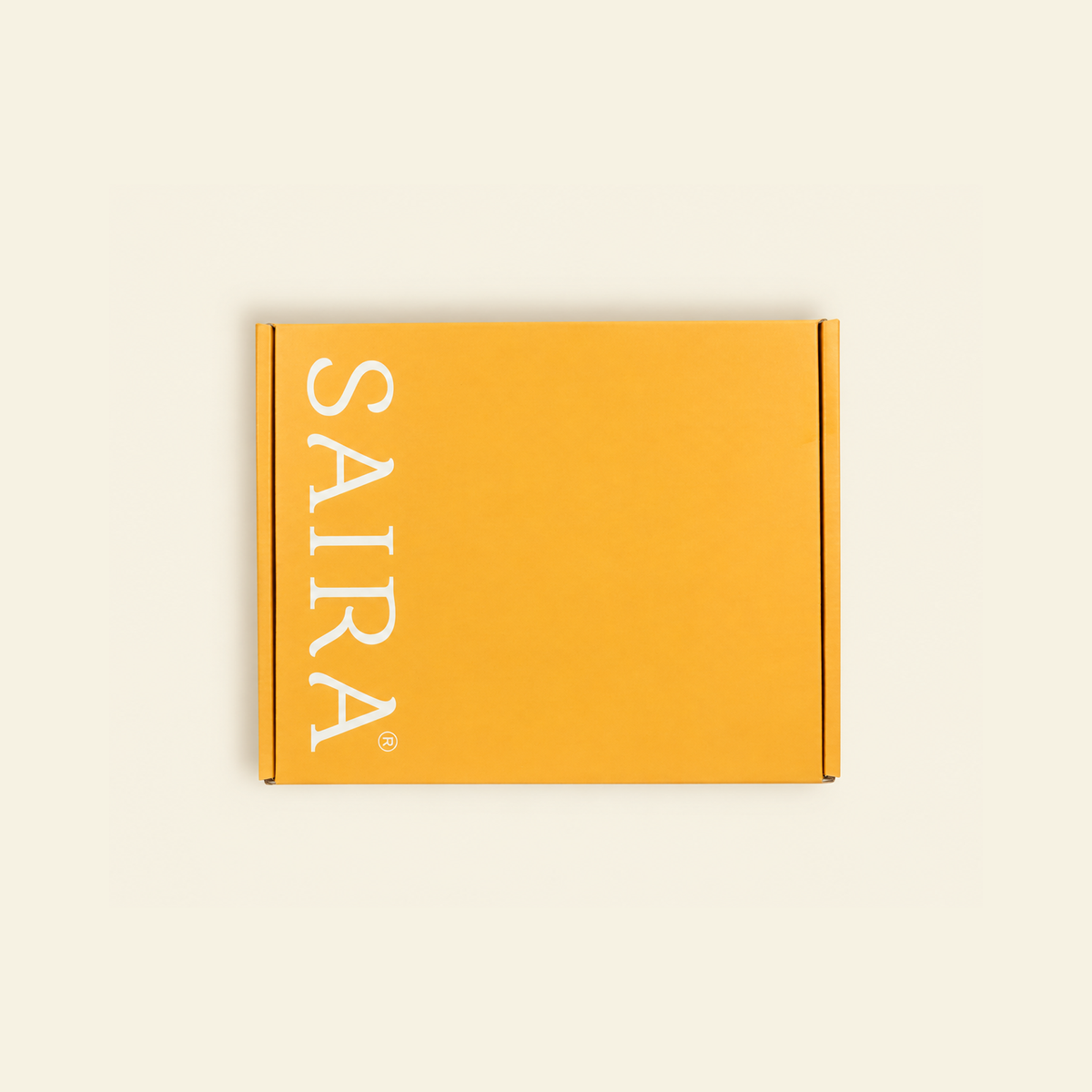 A mustard yellow cardboard box with the brand name 'SAIRA' printed vertically in white serif lettering on the left side, set against a solid off-white background.