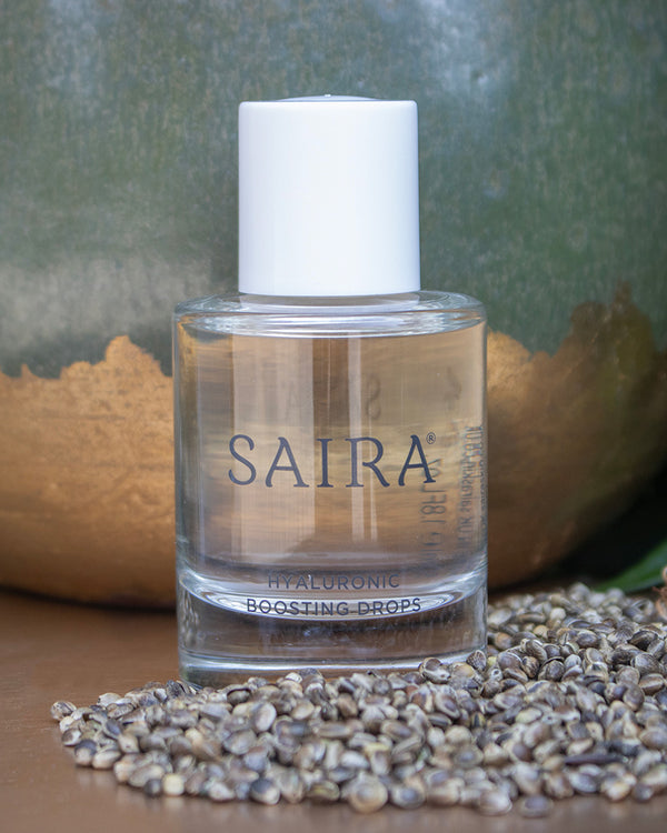 A bottle of Saira Hyaluronic Boosting Drops with a white cap and clear glass bottle showing the golden liquid inside, set on a wooden surface with sesame seeds in the foreground and a textured green and gold background.
