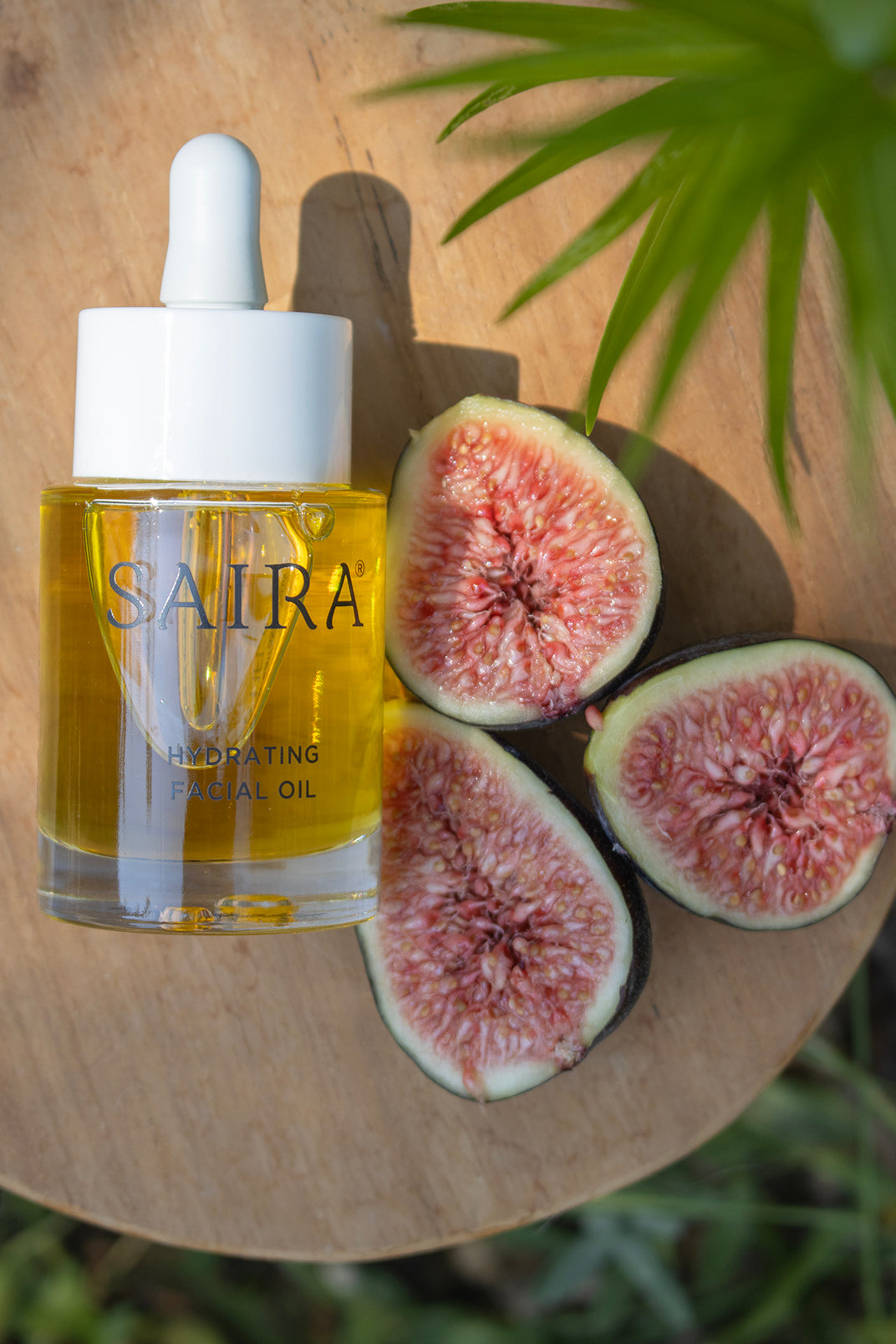SAIRA Hydrating Facial Oil in a clear glass dropper bottle with a white cap, styled on a wooden board next to three halved figs and green palm leaves.