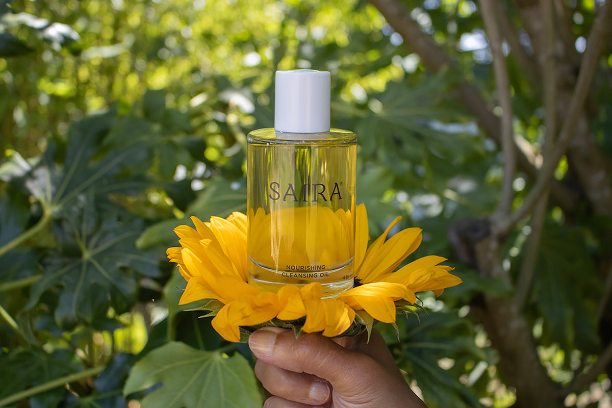 A hand holds a clear glass bottle of SAIRA Nourishing Cleansing Oil, positioned over a bright yellow sunflower, with a lush green garden background.