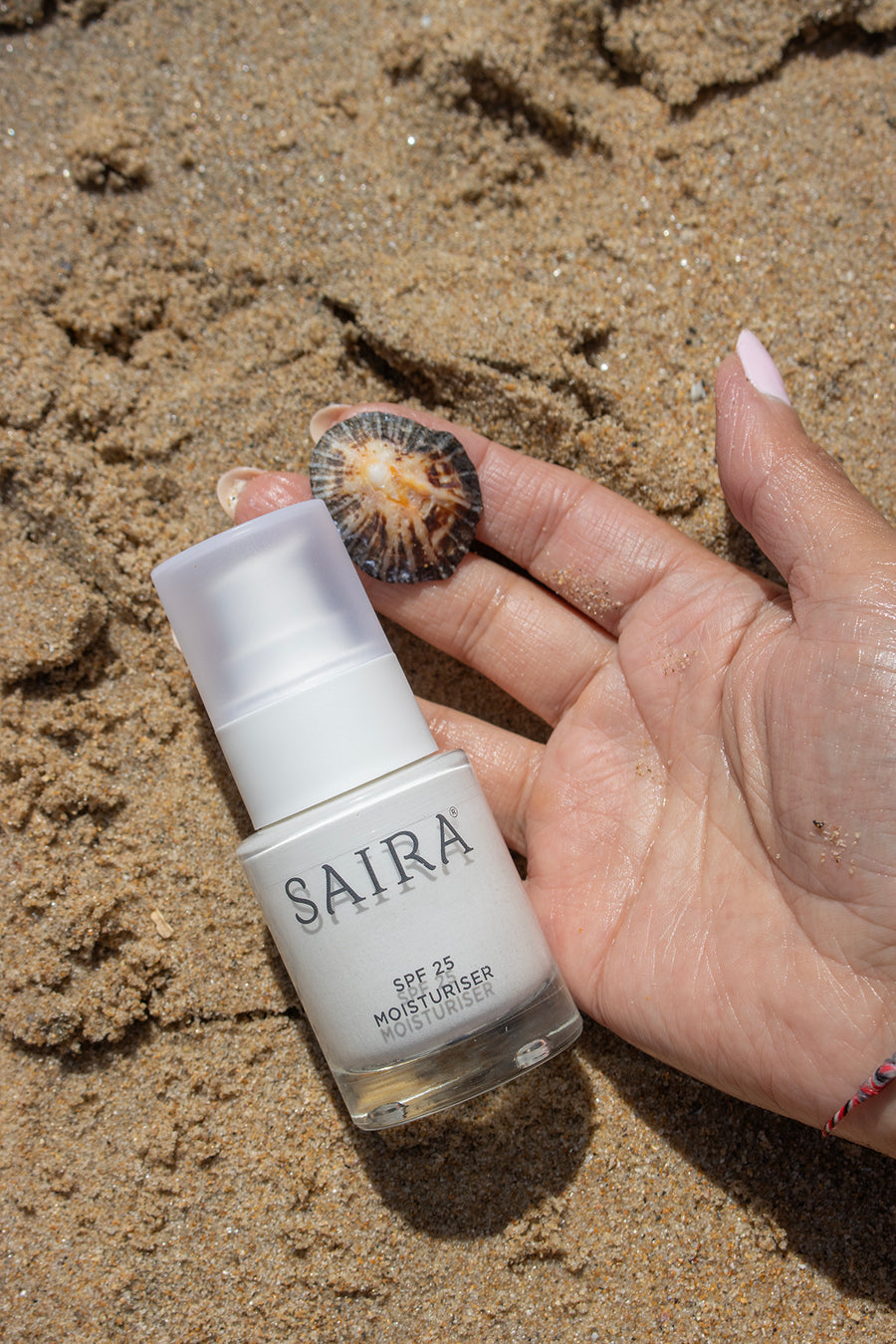 A hand holding a bottle of SAIRA SPF 25 Moisturiser on a sandy beach background.