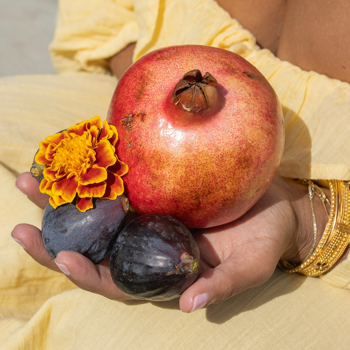 The hand of a woman wearing a yellow dress and a gold bangle cradles a pomegranate, some figs and a flower.