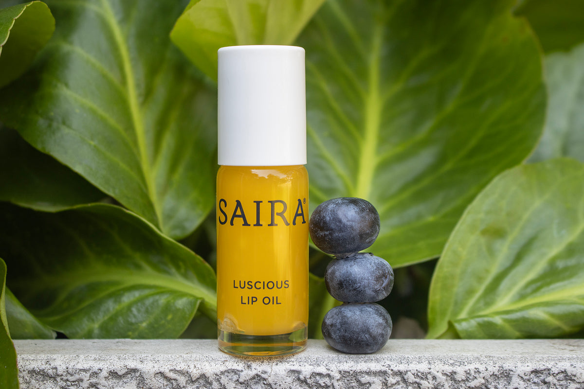 A bottle of SAIRA Luscious Lip Oil with a white cap stands on a stone surface, flanked by three blueberries and large green leaves in the background.