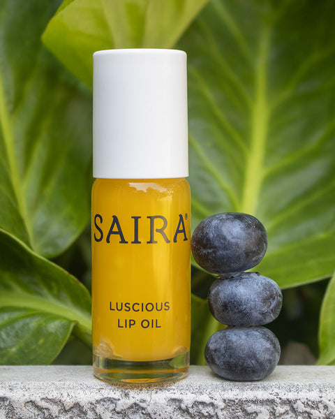 A bottle of Saira Luscious Lip Oil with a white cap and yellow-orange liquid, standing on a stone surface next to three blueberries in front of large green leaves.