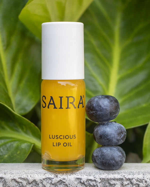 A bottle of Saira Luscious Lip Oil with a white cap and yellow-orange liquid, standing on a stone surface next to three blueberries in front of large green leaves.