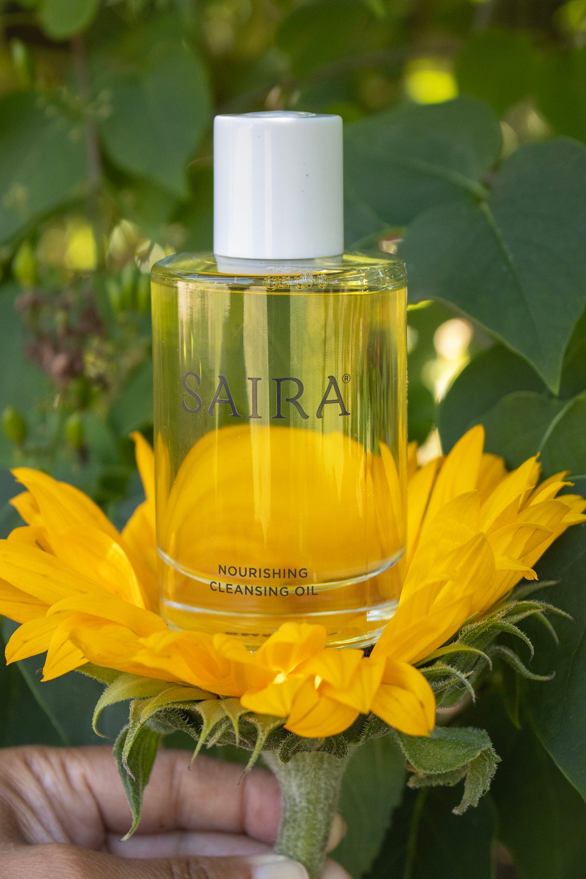 A bottle of SAIRA Nourishing Cleansing Oil held in front of a large yellow sunflower, with green leaves and a hand visible at the bottom left.
