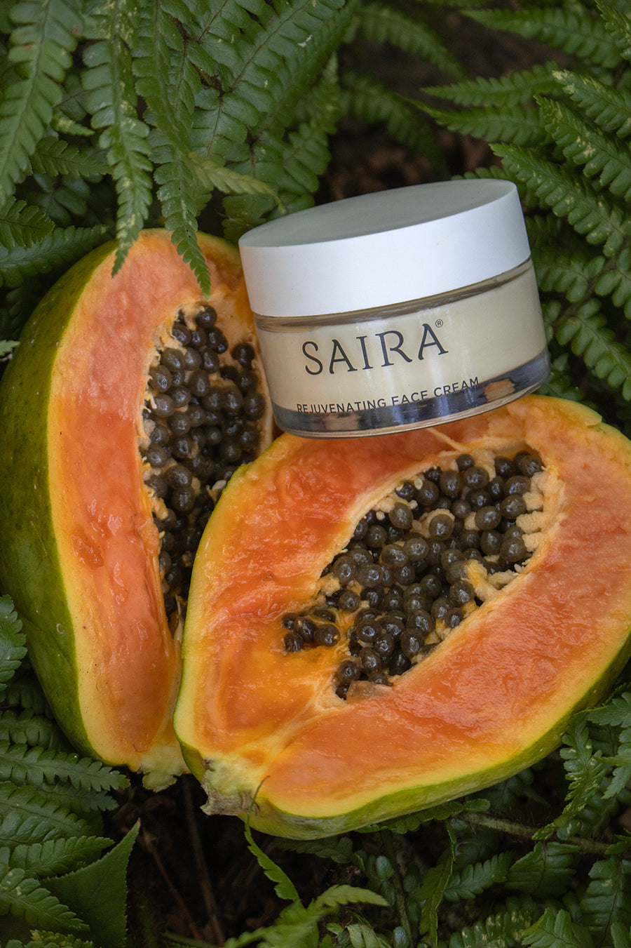 SAIRA Rejuvenating Face Cream glass jar placed on top of two halved papayas surrounded by green fern leaves.