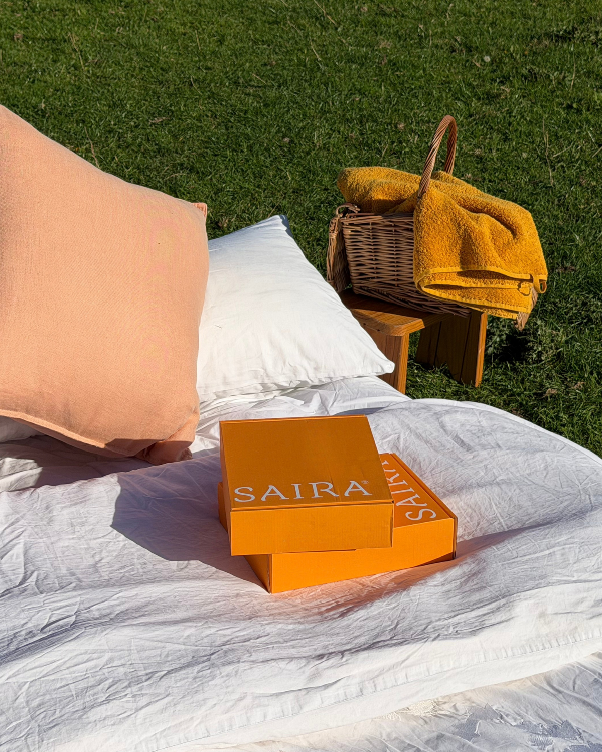 A styled outdoor picnic scene on a white blanket with a peach pillow, white pillow, and a folded mustard yellow towel in a wicker basket. Two orange Saira boxes are stacked on the blanket in the foreground, with green grass in the background.