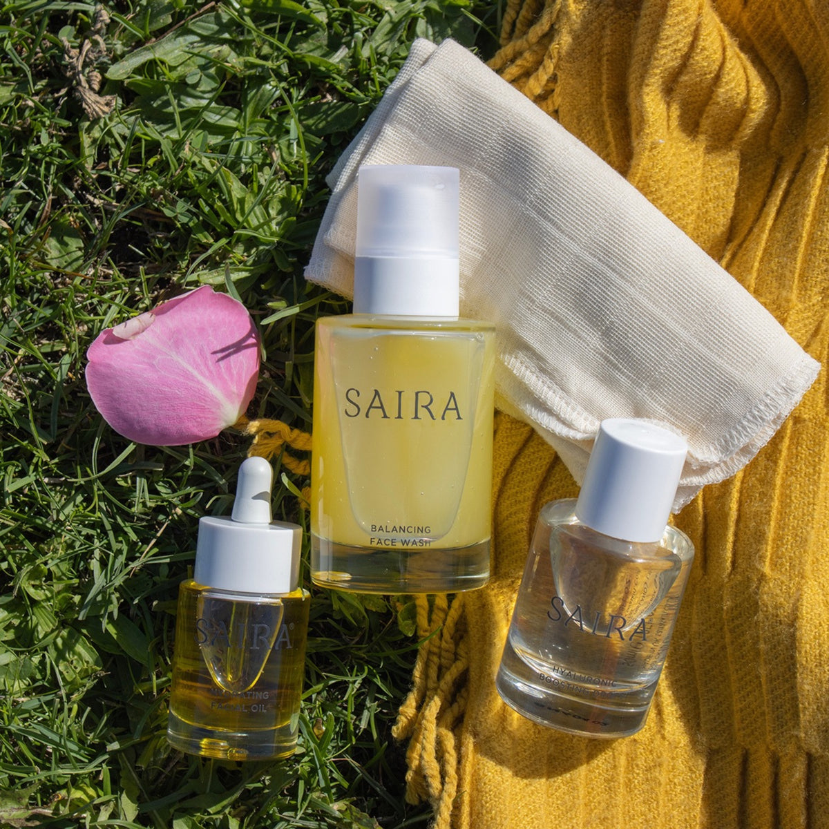 Three Saira skincare products arranged on green grass with a pink flower petal, a white cotton cloth, and a yellow knit blanket. The products include a balancing face wash bottle, a serum dropper bottle, and a clear serum bottle.