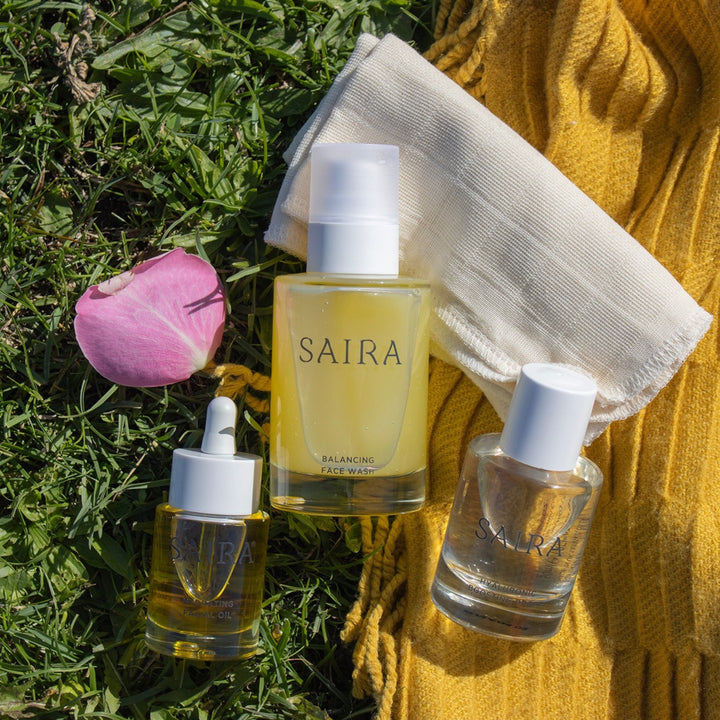 Three Saira skincare products arranged on green grass with a pink flower petal, a white cotton cloth, and a yellow knit blanket. The products include a balancing face wash bottle, a serum dropper bottle, and a clear serum bottle.