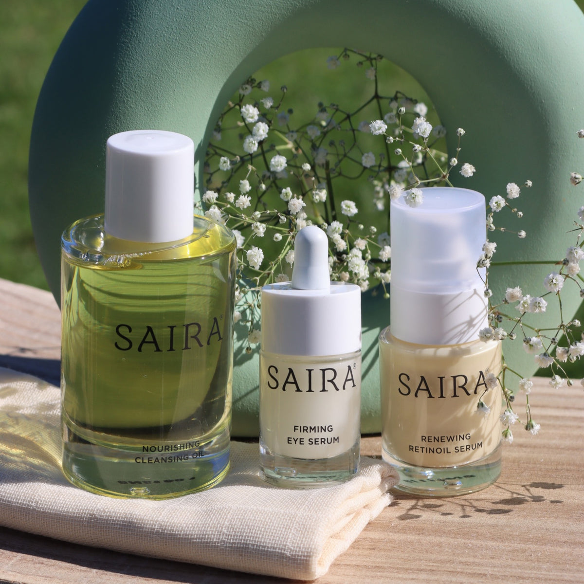 Three SAIRA skincare bottles and dropper bottles arranged on a folded cream cloth outdoors on a wooden surface, with a soft green arch and white baby's breath flowers in the background.