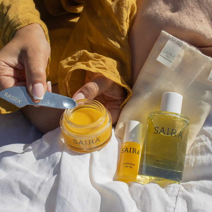 A person's hands holding a metal spatula over a jar of yellow cream, with Saira skincare products including a small bottle of lip oil, a larger bottle of cleansing oil, and a mesh pouch arranged on white fabric in warm natural light.