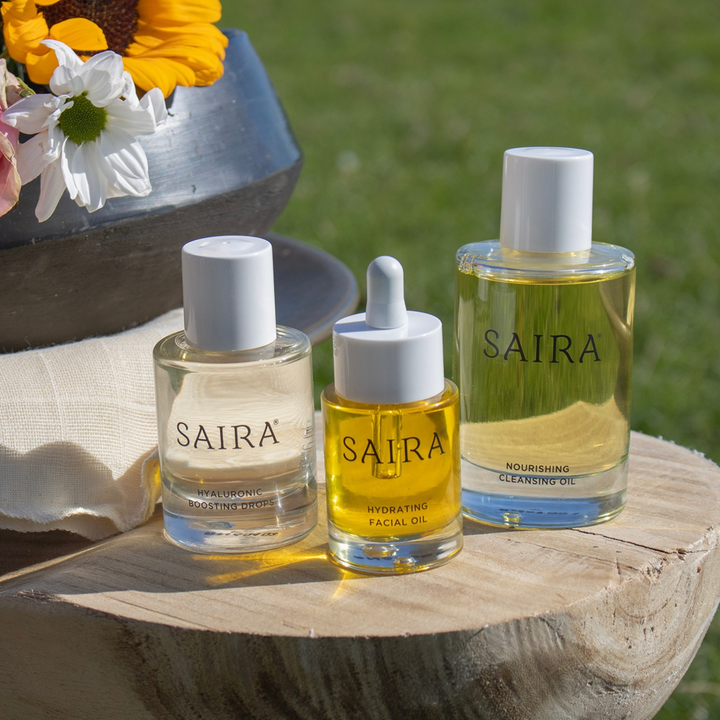 Three Saira facial care bottles arranged on a wooden surface outdoors, with flowers and a blurred green lawn in the background. The bottles contain clear and golden-yellow liquids.