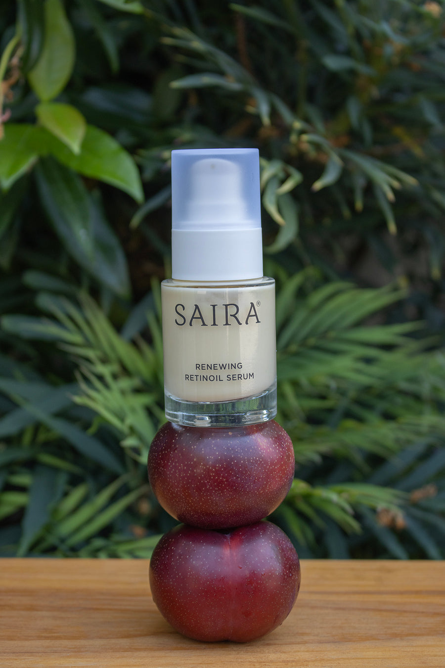 SAIRA Renewing Retinol Serum in a clear glass bottle with a white pump, stacked on top of two dark red plums on a wooden surface with a blurred green garden background.