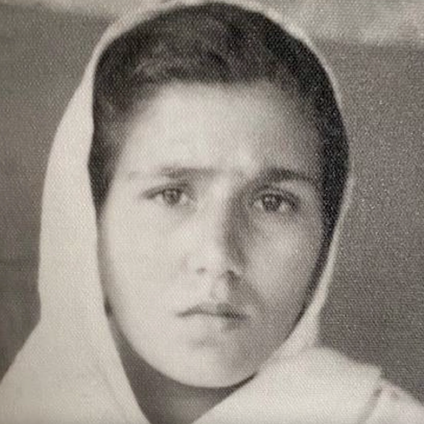 A black and white portrait of a young woman wearing a light-colored headscarf or hood, shown in a grainy, vintage-style photograph.