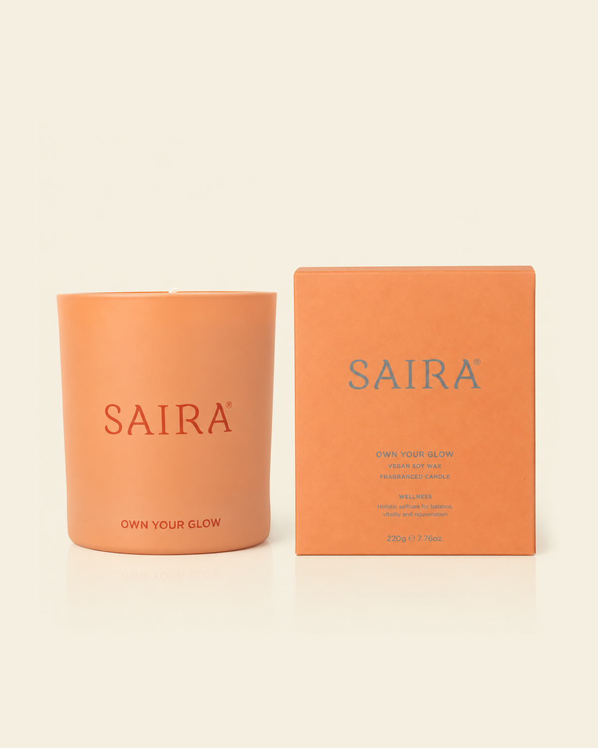 An orange candle in a matte orange glass vessel next to its matching orange packaging box, both featuring the brand name 'SAIRA' in blue, set against a solid cream background.