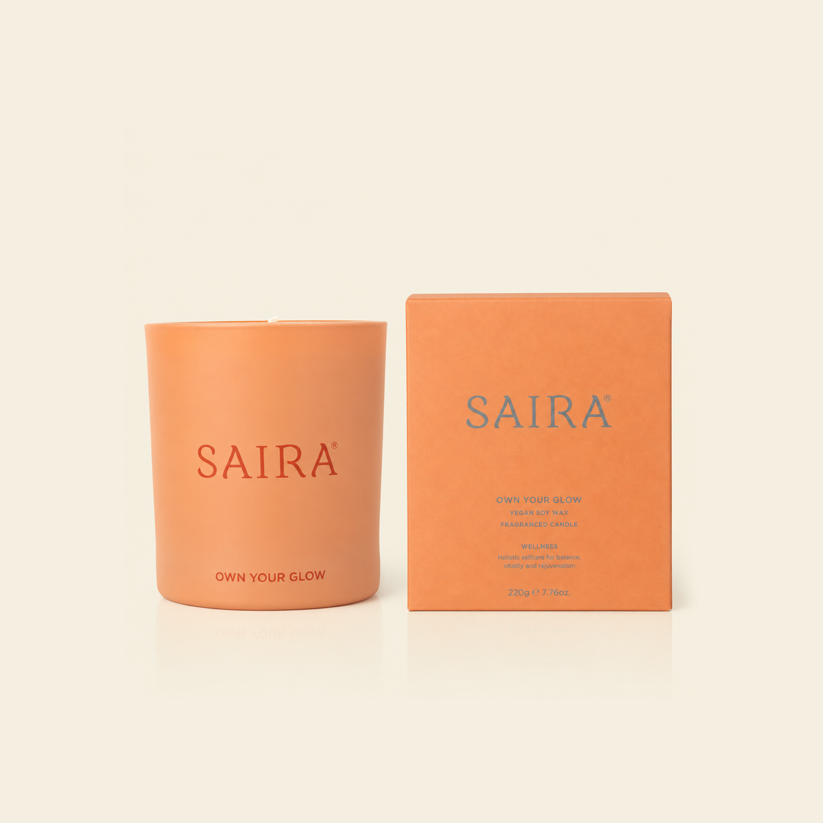 An orange candle in a matte orange glass vessel next to its matching orange packaging box, both featuring the brand name 'SAIRA' in blue, set against a solid cream background.