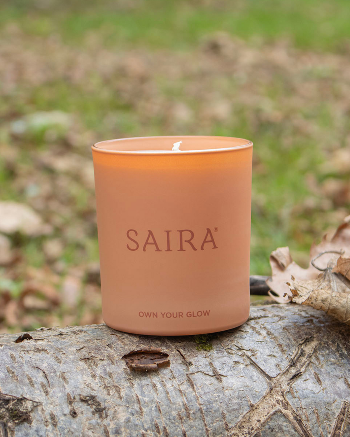 A lit Saira scented candle in a matte terracotta-colored glass jar, resting on a weathered log outdoors with a blurred natural background of grass and dry leaves.