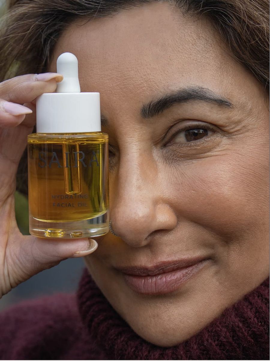A close-up lifestyle shot of Saira holding a bottle of SAIRA Hydrating Facial Oil next to her face. The bottle is filled with golden-yellow oil and has a white dropper cap, set against a soft-focus background of her skin and a burgundy knit sweater.