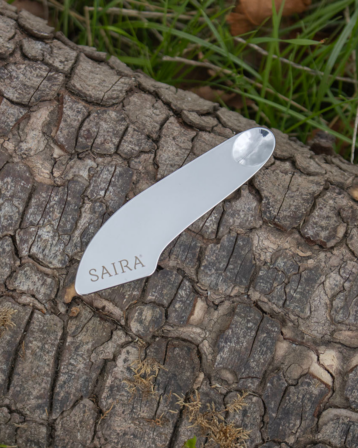 A silver metal spatula with the brand name 'SAIRA' engraved on it, resting on a textured tree trunk with green grass in the background.