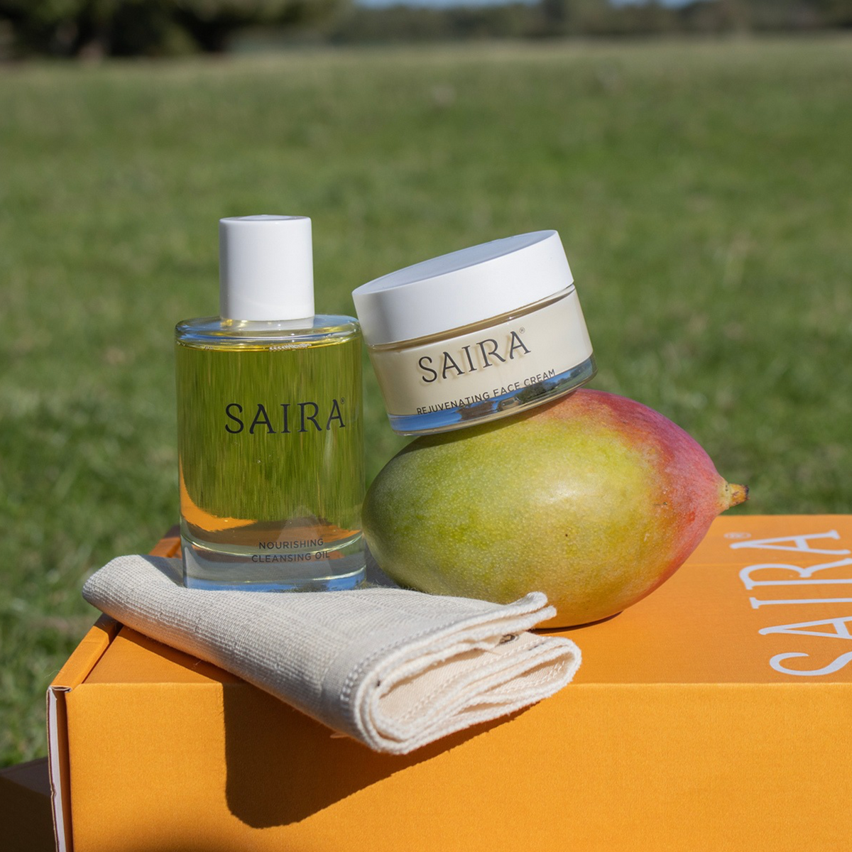 SAIRA skincare products displayed outdoors on an orange box: a clear glass bottle with yellow liquid and white cap, a white-lidded cream jar labelled SAIRA, a mango, and a folded cream cloth. A blurred green field is in the background.