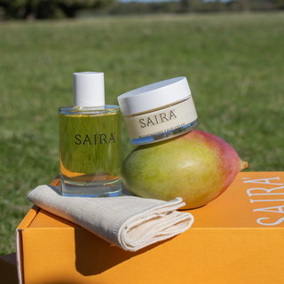 SAIRA skincare products displayed outdoors on an orange box: a clear glass bottle with yellow liquid and white cap, a white-lidded cream jar labelled SAIRA, a mango, and a folded cream cloth. A blurred green field is in the background.
