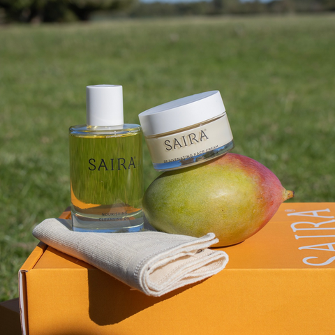 SAIRA skincare products displayed outdoors on an orange box: a clear glass bottle with yellow liquid and white cap, a white-lidded cream jar labelled SAIRA, a mango, and a folded cream cloth. A blurred green field is in the background.