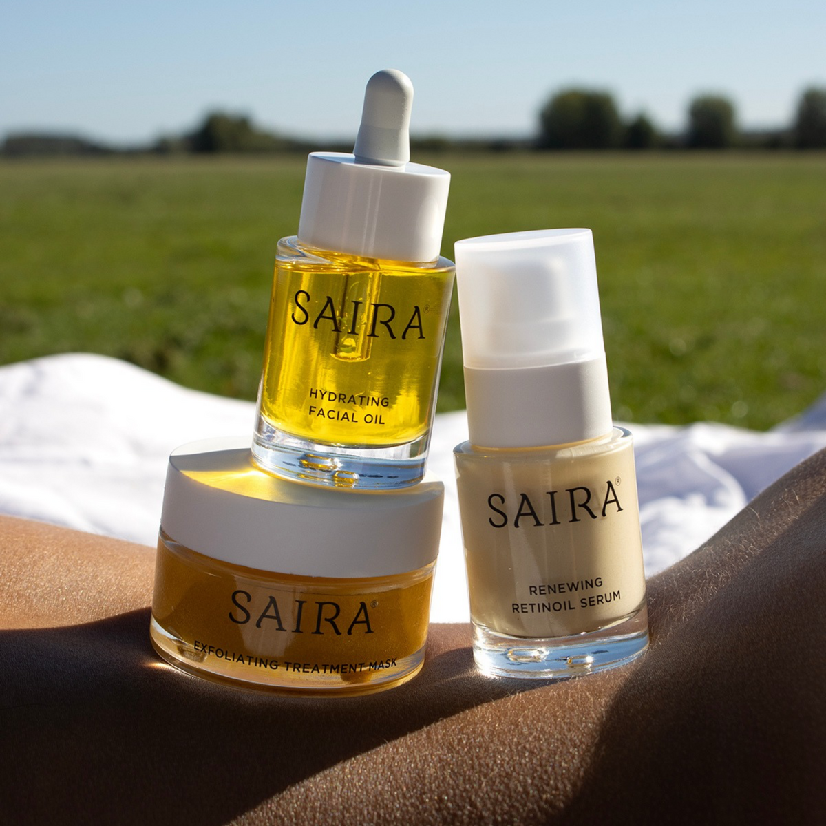 Three Saira skincare products arranged on a person's lap outdoors: a jar of exfoliating treatment cream, a dropper bottle of hydrating facial oil, and a pump bottle of renewing retinoil serum. The background shows a blurred green field and blue sky.