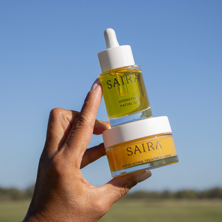 A hand holds two Saira skincare products against a bright blue sky background: a small glass dropper bottle of hydrating facial oil and a jar of exfoliating treatment balm.