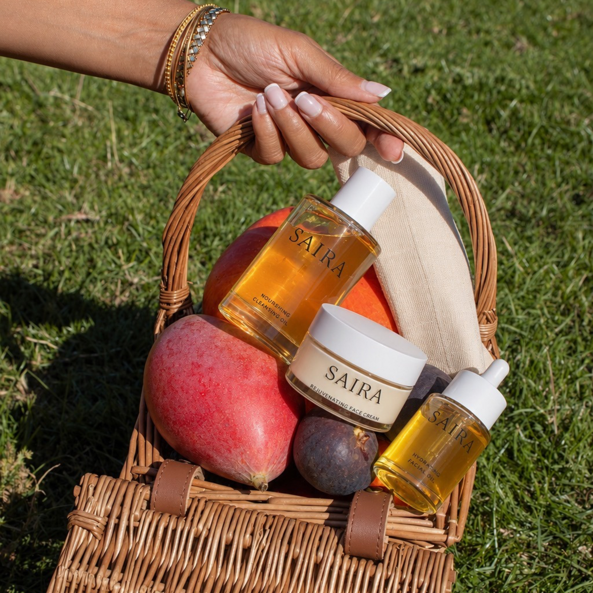 A hand holds a wicker picnic basket outdoors on grass, containing two Saira skincare bottles and a cream jar alongside fruit and a beige cloth.