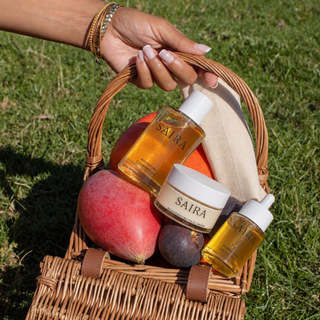 A hand holds a wicker picnic basket outdoors on grass, containing two Saira skincare bottles and a cream jar alongside fruit and a beige cloth.