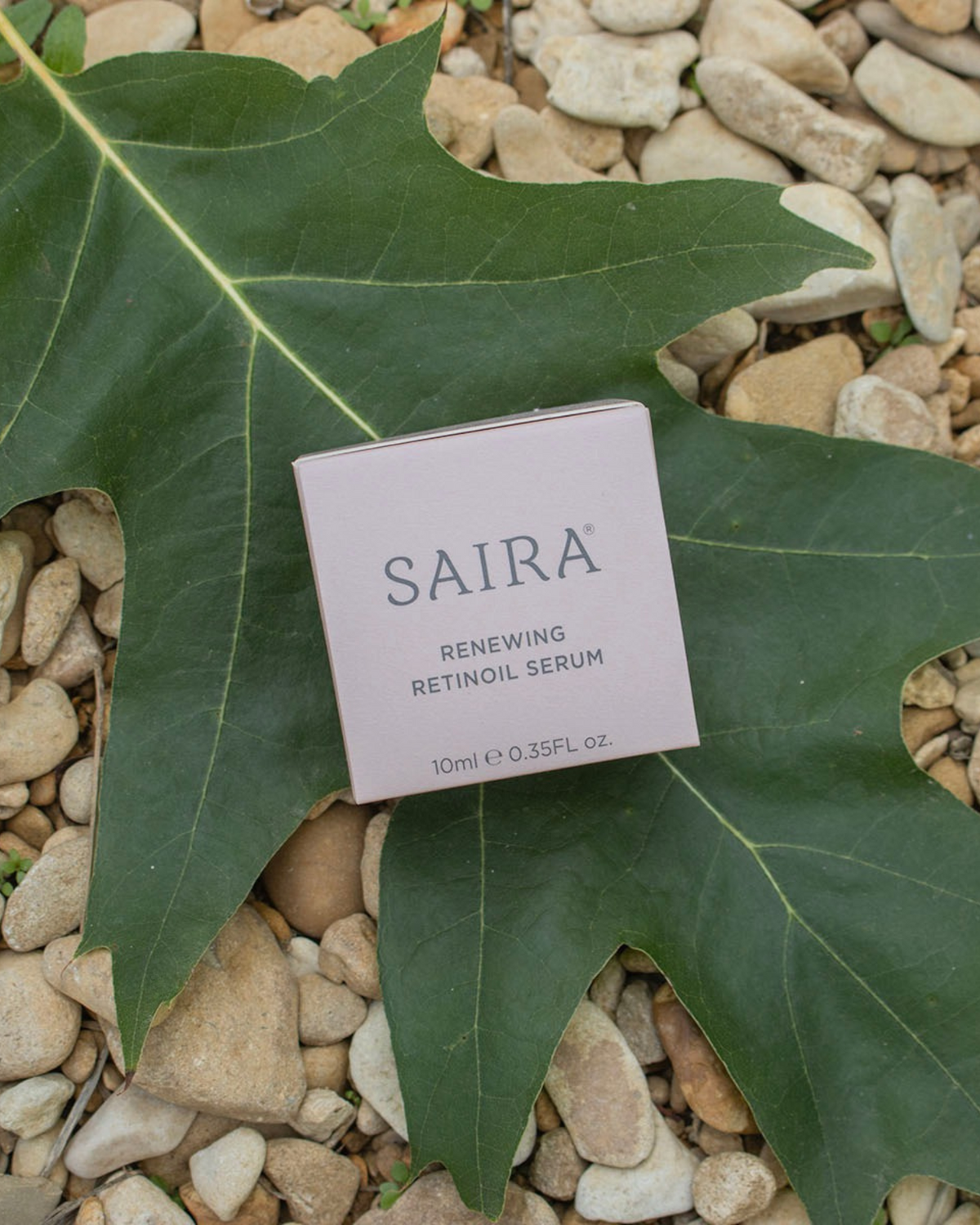 A square box of Saira Renewing Retinol Serum lies on a large green leaf over a bed of small beige pebbles. The box is pale pink with dark gray text.