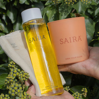 A person holds a clear bottle of Saira body oil with yellow liquid, a peach-colored Saira candle, and a beige cloth against a background of green leaves and small yellow flowers.