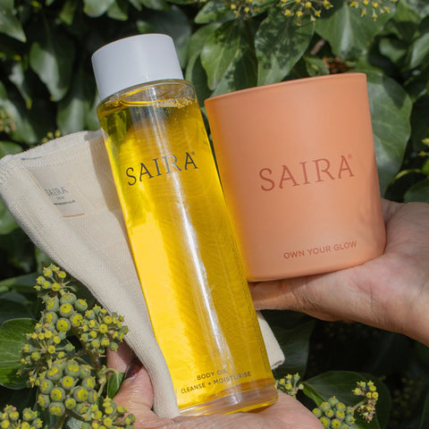 A person holds a clear bottle of Saira body oil with yellow liquid, a peach-colored Saira candle, and a beige cloth against a background of green leaves and small yellow flowers.