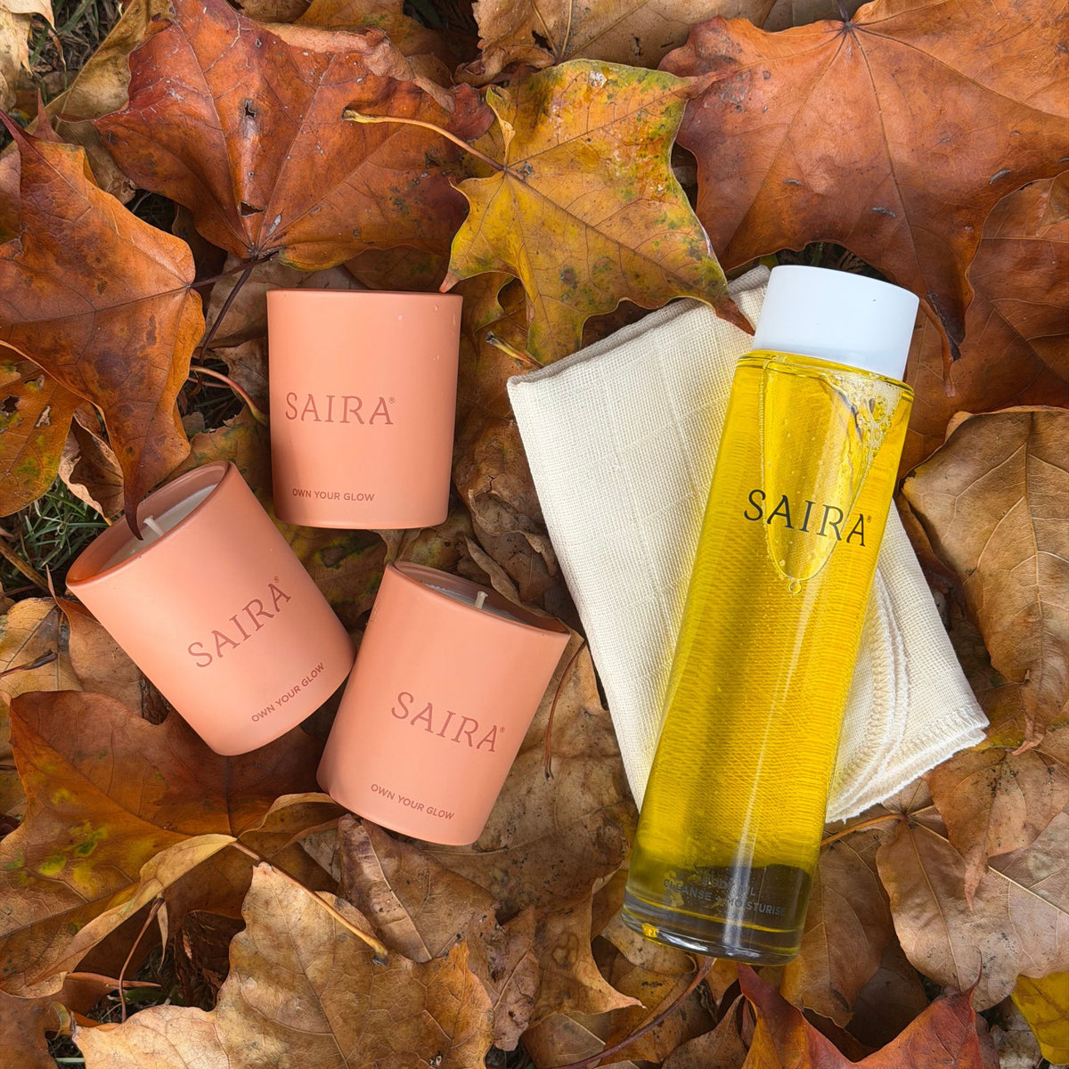 Five Saira skincare products arranged on a bed of autumn leaves: two peach-coloured votive candles, one clear bottle filled with yellow oil, and a folded cream cloth. The products are branded with the Saira name in black lettering.