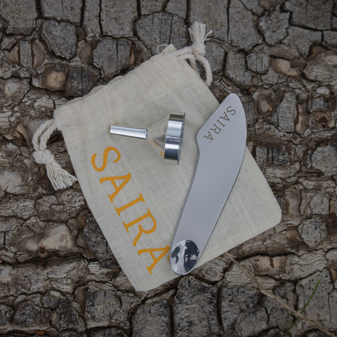 A small stainless steel funnel and stainless steel facial spatula sitting on a small cotton drawstring pouch printed with the SAIRA logo in safffron yellow, placed on a textured tree bark background.